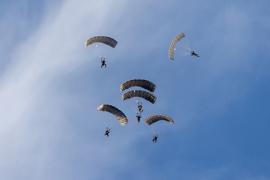 Six Exercise Rising Gains 1‑26 parachutists descending through a blue sky with open desert sand coloured rectangular parachutes, including two in a stacked formation.